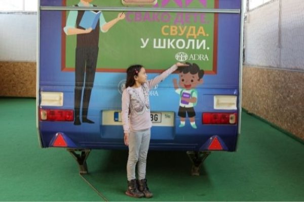 Eine junge Schülerin steht vor einem mobilen Schulbus und zeigt mit der linken Hand auf das ADRA Logo
