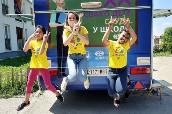 Drei fröhliche Schulmädchen springen vor der mobilen Schule in die Luft und zeigen mit beiden ihrer Händen das Peace-Zeichen
