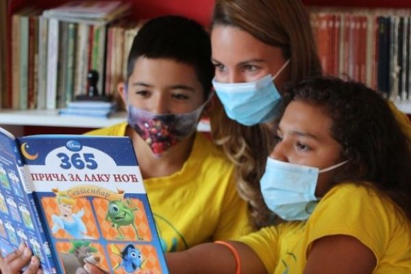 zwei Schulkinder und eine Betreuerin tragen Mund-und-Nasen-Schutz-Masken und lesen gemeinsam aus einem Disney-Buch vor einem Regal mit vielen Büchern und einem Stempel