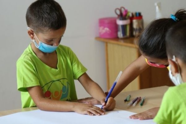 Drei junge Schüler sitzen an einem Tisch, tragen Mund-Schutz-Bedeckung und malen gemeinsam auf einem Papier