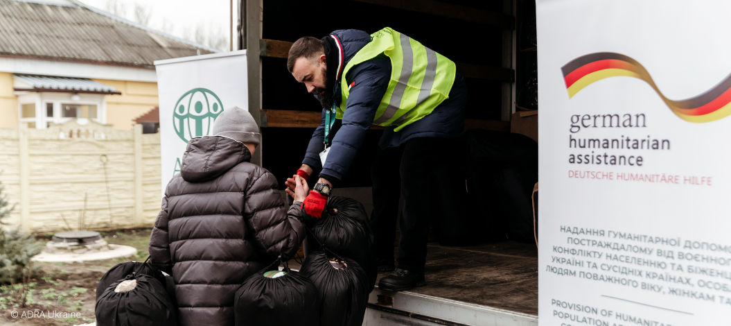 Ein ADRA-Mitarbeiter aus der Ukraine zeigt Mitgefühl, indem er eine Vielzahl von Lebensmitteln und Hilfsgütern aus einem Transporter an eine Ukrainerin verteilt, die vom Krieg schwer getroffen wurde.