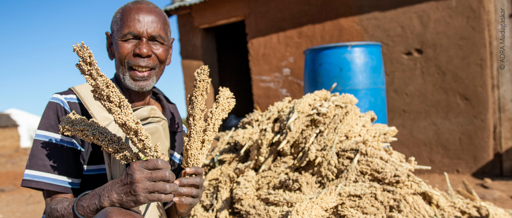 Ein freundlich lächelnder Mann aus Madagaskar sitzt im Freien und hält Getreide in den Händen. Im Hintergrund erstreckt sich ein Berg voller Getreide