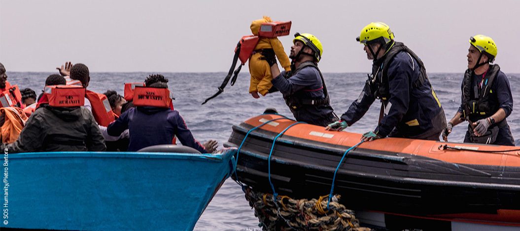 Boot mit Geflüchteten neben Rettungsboot von SOS Humanity – Kleinkind wird ins Rettungsboot gehoben. © SOS Humanity/Pietro Bertora