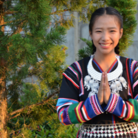 Ein fröhlich strahlendes Mädchen in einem bunten traditionellen Kleid hält die Hände vor einer Tanne zusammen