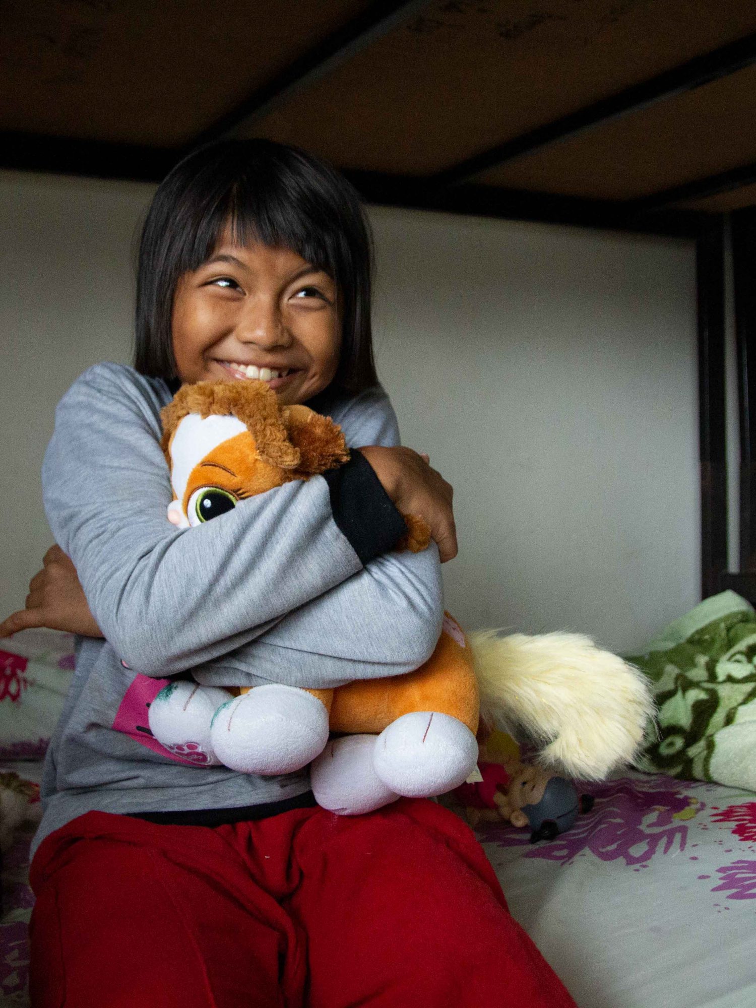 Ein lächelndes Mädchen sitzt auf dem Bett und hält ein Kuscheltier ganz fest im Arm