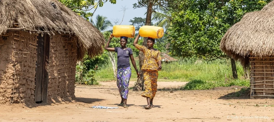 Zwei Frauen tragen Wasserkanister durch ein Dorf in der Demokratischen Republik Kongo, um ihre Wasserversorgung und Lebensgrundlagen zu sichern.