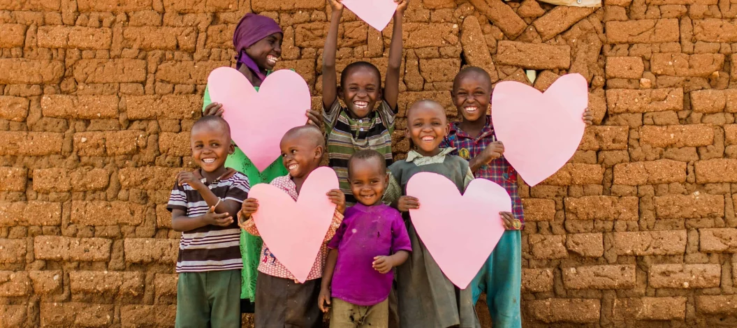 DZI Siegel ADRA Deutschland 2021 - Lachende Kinder in Kenia halten große rosa Herzen vor einer Mauer aus Lehmziegeln