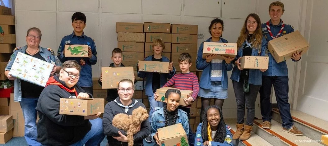 Kinder, Jugendliche und Erwachsene stehen gemeinsam in einem Raum und halten Geschenkpakete der Aktion ›Kinder helfen Kindern‹ in den Händen. Im Hintergrund sind viele weitere Pakete gestapelt. Die Gruppe lächelt in die Kamera