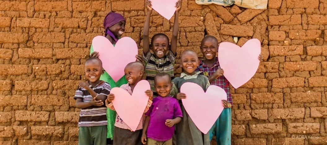 Lachende Kinder in Kenia halten große rosa Herzen vor einer Mauer aus Lehmziegeln