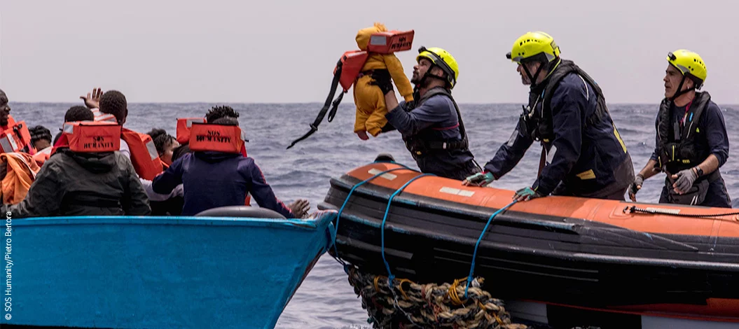 Boot mit Geflüchteten neben Rettungsboot von SOS Humanity – Kleinkind wird ins Rettungsboot gehoben. © SOS Humanity/Pietro Bertora