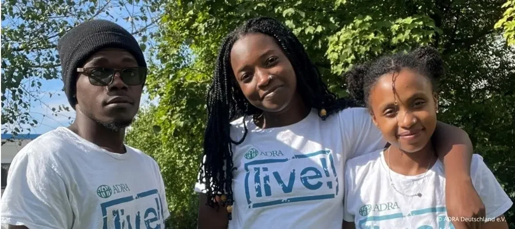 Drei junge Personen stehen draußen im Grünen vor Bäumen. Alle tragen weiße ADRAlive! T-Shirts. Die Person links trägt eine Mütze und Brille, die mittlere Person lächelt und hat lange geflochtene Haare, und die Person rechts lehnt sich an die mittlere und lächelt ebenfalls.