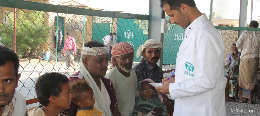 ADRA-Mitarbeiter im weißen Kittel spricht mit wartenden Männern und Kindern in einer Einrichtung im Jemen