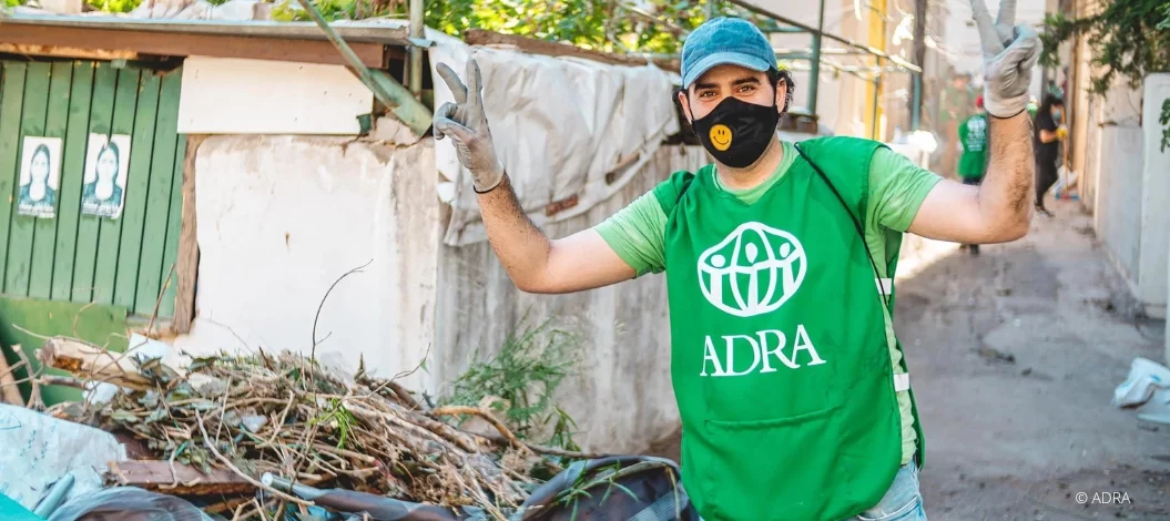 ADRA-Mitarbeiter in grüner Weste, blauer Basecap, Handschuhen und Smiley-Mundschutz hebt die Arme und zeigt das Peace-Zeichen