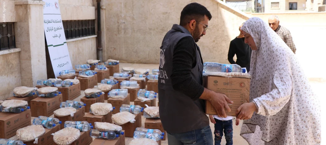 ADRA-Helfer überreicht syrischer Frau Hilfspaket mit Wasser und Fladenbrot – umgeben von weiteren gepackten Kartons
