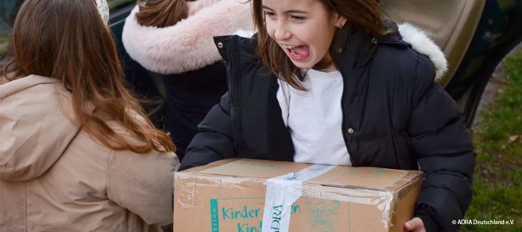 Mädchen trägt ein Hilfspaket mit dem Logo der Aktion „Kinder helfen Kindern!“ und lächelt freudig.