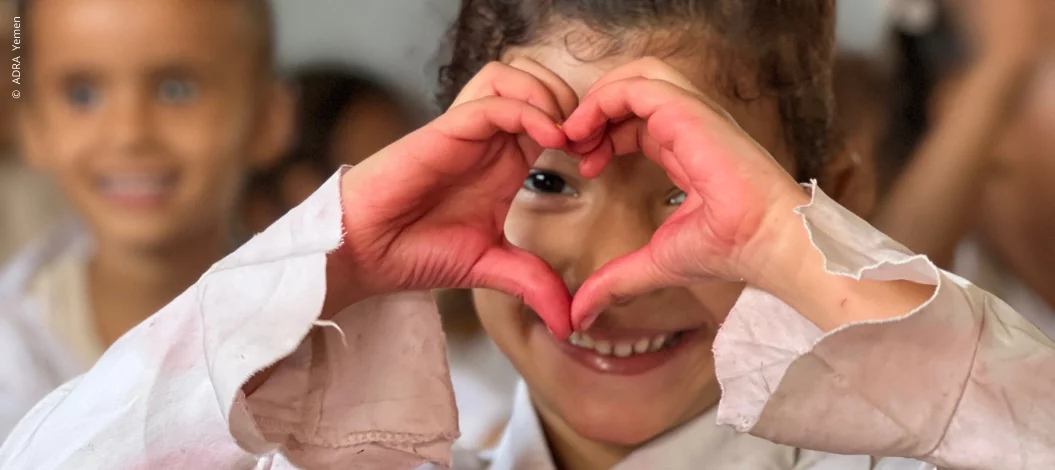 Mädchen in jemenitischer Schulklasse formt mit rot bemalten Händen ein Herz vor dem Gesicht und blickt hindurch