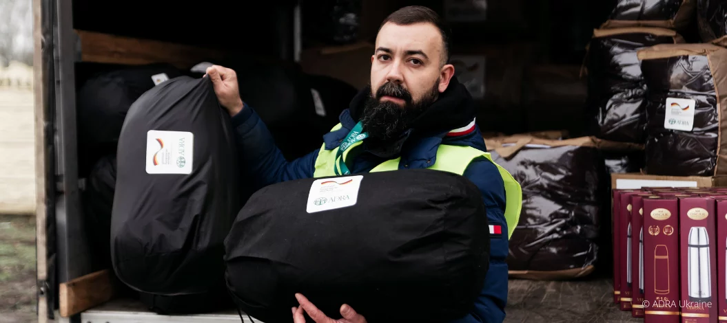 Ein ADRA-Mitarbeiter in gelber Warnweste hält zwei schwarze Hilfspakete mit ADRA-Logo vor einem Transporter voller weiterer Hilfsgüter