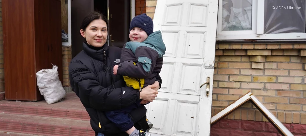 Junge Mutter aus der Ukraine hält ihren Sohn vor zerstörtem Haus – benötigt Winterhilfe