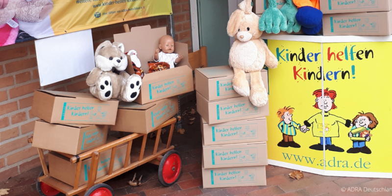 Kartons der ADRA-Aktion „Kinder helfen Kindern!“ mit Spielzeug und Plüschtieren auf Bollerwagen und neben Werbeschild