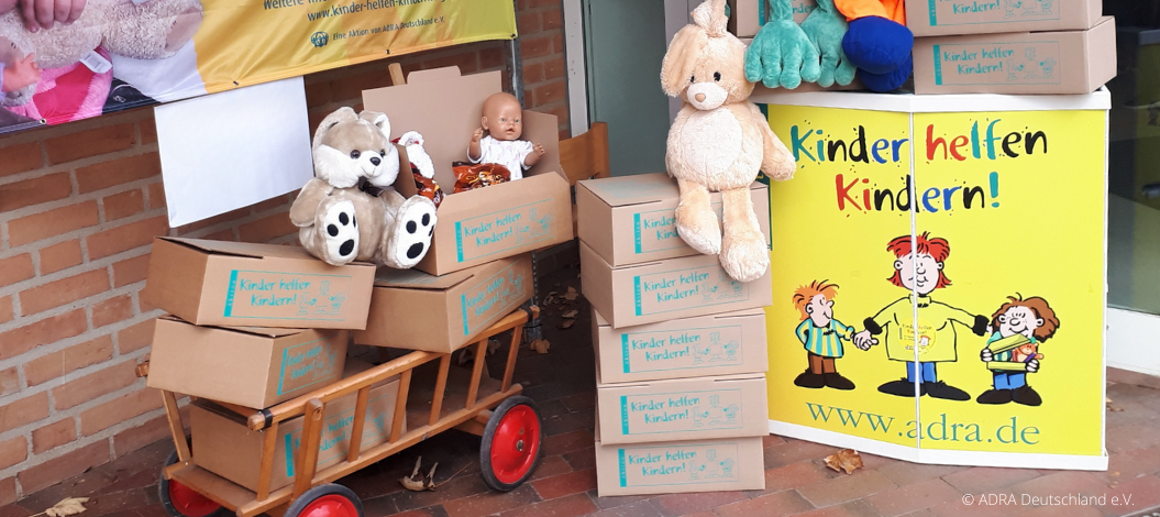 Kartons der ADRA-Aktion „Kinder helfen Kindern!“ mit Spielzeug und Plüschtieren auf Bollerwagen und neben Werbeschild